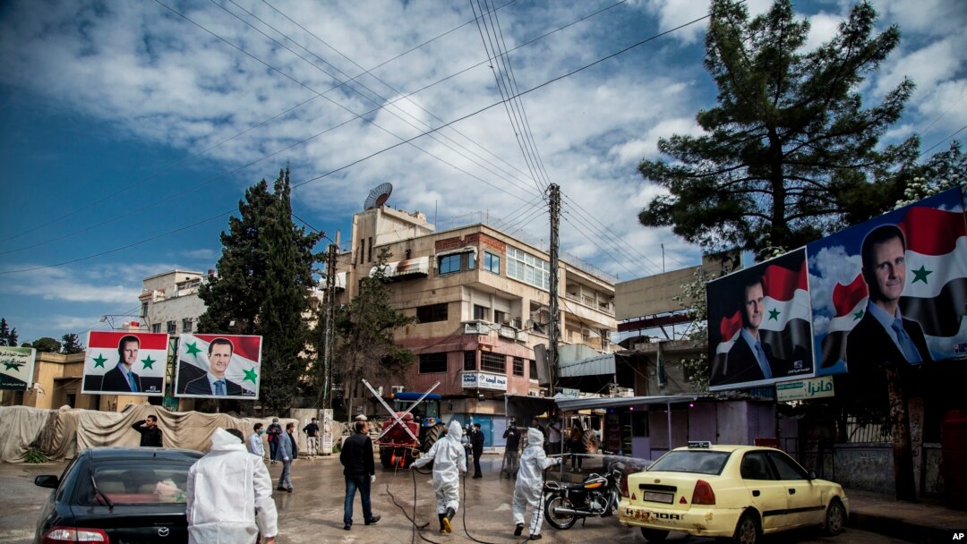 FILE - Workers spray disinfectant to prevent the spread of the coronavirus, on a street lined with billboards showing Syrian President Bashar al-Assad, in Qamishli, Syria, March 24, 2020.