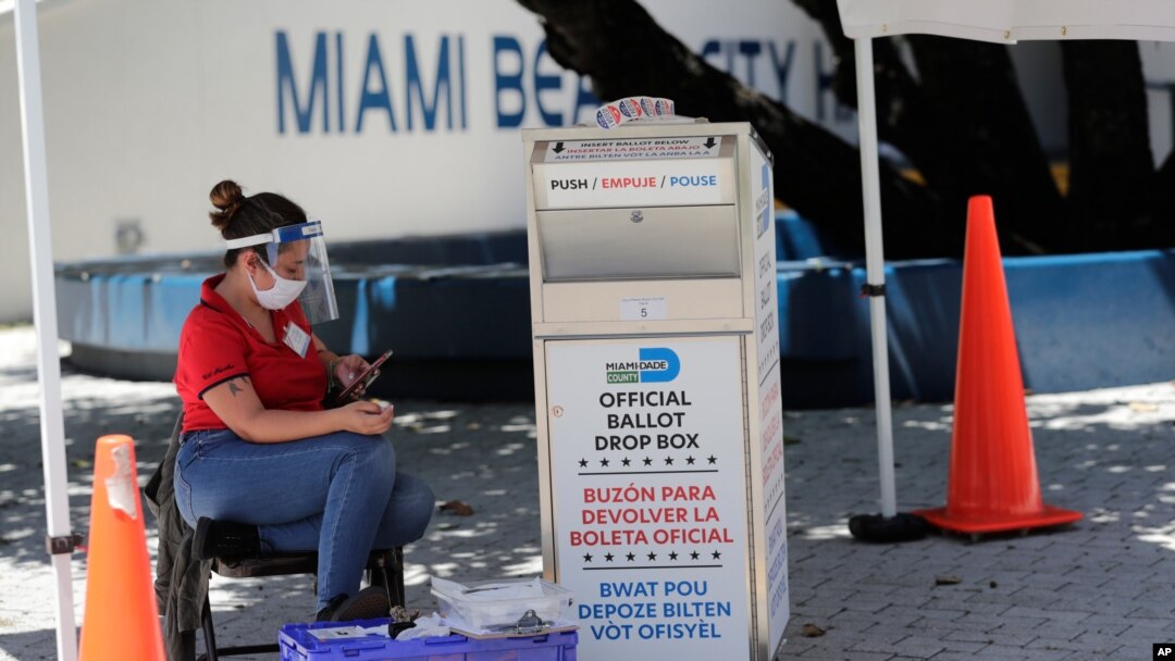 A poll worker wears personal protective equipment as she monitors a ballot drop box for mail-in ballots outside of a polling station during early voting, Aug. 7, 2020, in Miami Beach, Florida. 