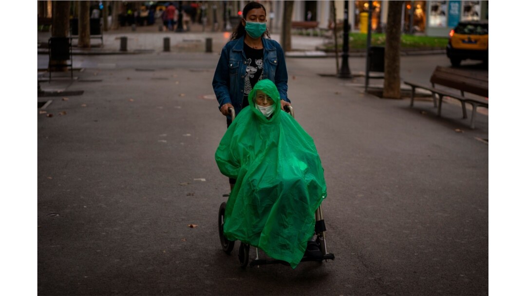 A man wearing a face mask and rain cover is pushed in a wheelchair along a boulevard in Barcelona, Spain, Sept. 9, 2020.