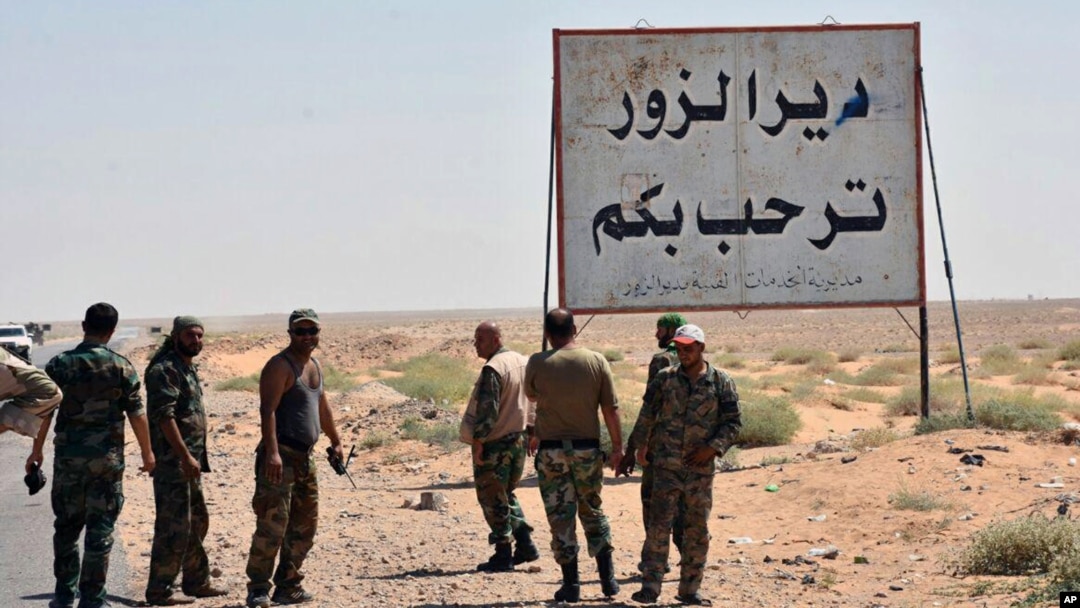 FILE - This photo released Sept 3, 2017 by the Syrian official news agency SANA, shows Syrian troops and pro-government gunmen standing next to a placard in Arabic which reads, "Deir el-Zour welcomes you," in the eastern city of Deir el-Zour, Syria. 